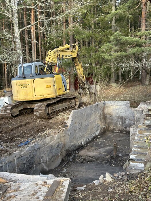 Gul grävmaskin vid riven husgrund omgiven av skog, med trasig betong runt en byggplats.