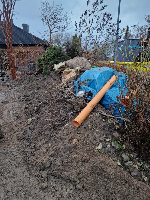 Hög med jord och vegetation framför ett hus, blå presenning och orange rör synliga i trädgården.