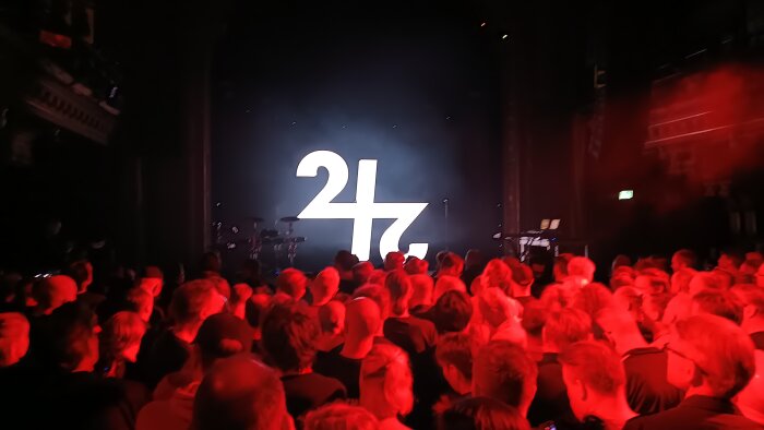 Stort publikhav i röd belysning framför en scen med vit text "24" och instrument, på en konsert eller evenemang i en mörk sal.