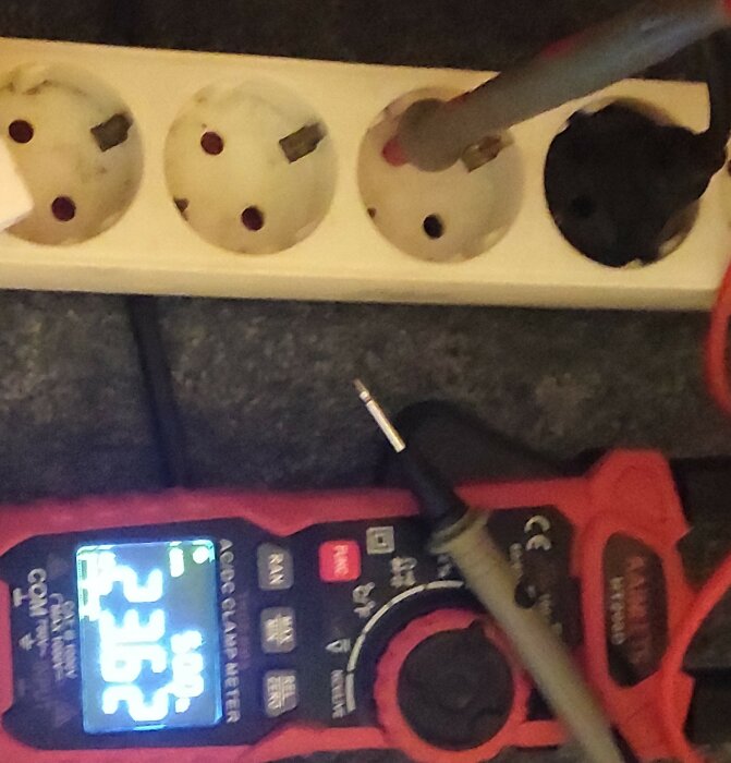 Multimeter med ansluten prob visar 23,6V AC; proben är placerad i ett grenuttag, medan den andra är i luften.