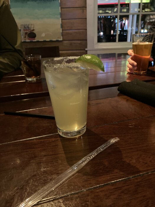 Ett glas margarita med limeklyfta och ett glas öl bakom, stående på ett träbord i en bar.