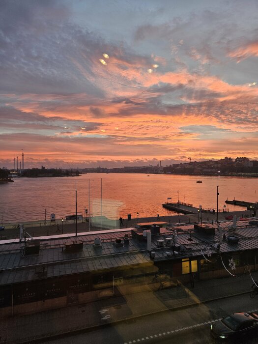 Solnedgång över en stad vid vattnet med moln i rosa och orange toner, och en kaj med människor i förgrunden.