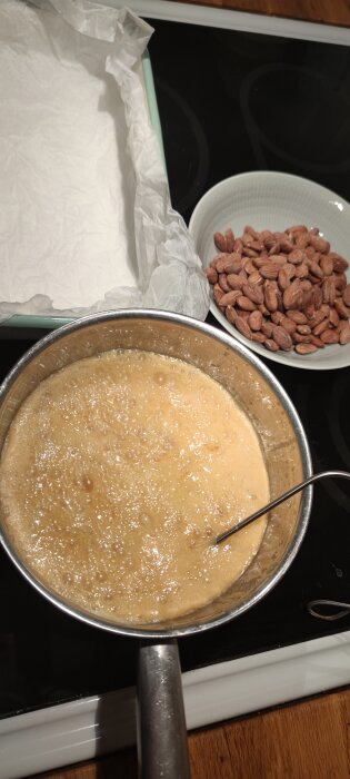 En gryta med bubblande gräddkola, en skål med mandlar och en bakform fodrad med bakplåtspapper på en spis.