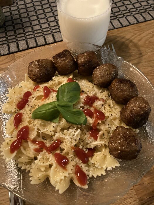 Tallrik med fjärilspasta, köttbullar, ketchup, riven ost och basilikablad. Glas med mjölk i bakgrunden.