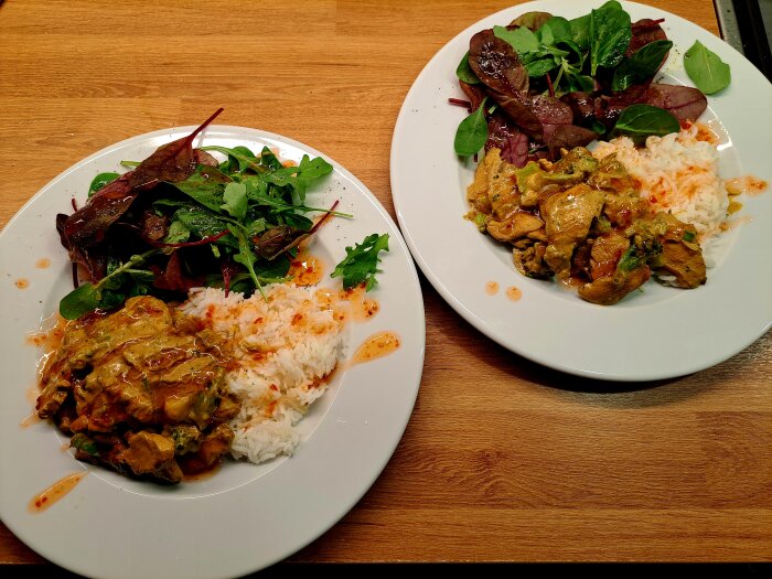 Två tallrikar med kyckling i currysås, ris och sallad på ett träbord.