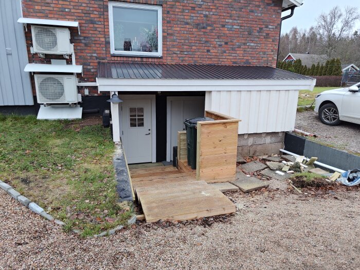 Trätrapp och paneltak leder ner till en källaringång vid ett hus. Luftkonditioneringsenheter monterade på fasaden. Grus och gräs omger området.