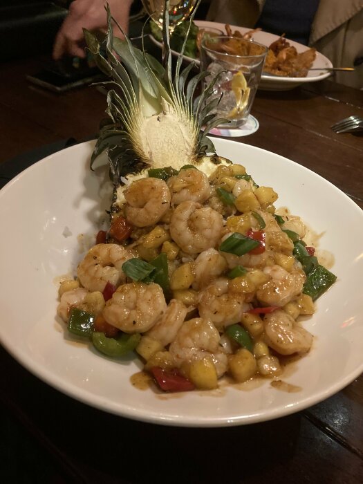 Räkrätt serverad i ananasskål med grönsaker, kryddad sås, och ananasblad som dekoration på en restaurangbord.