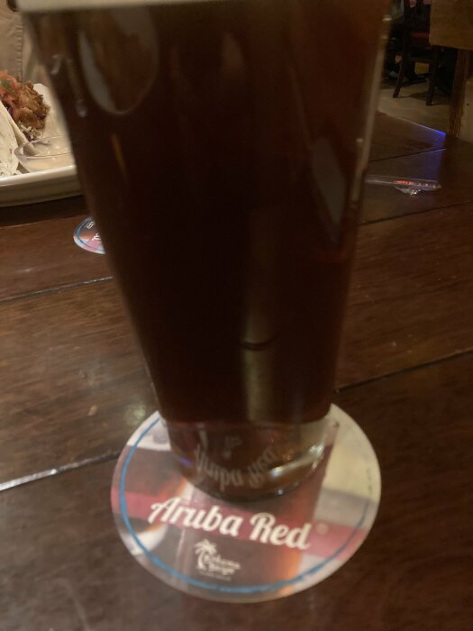 Mörk öl i ett glas på ett bord, placerad på en ölunderlägg märkt "Aruba Red". I bakgrunden syns en del av en tallrik med mat.