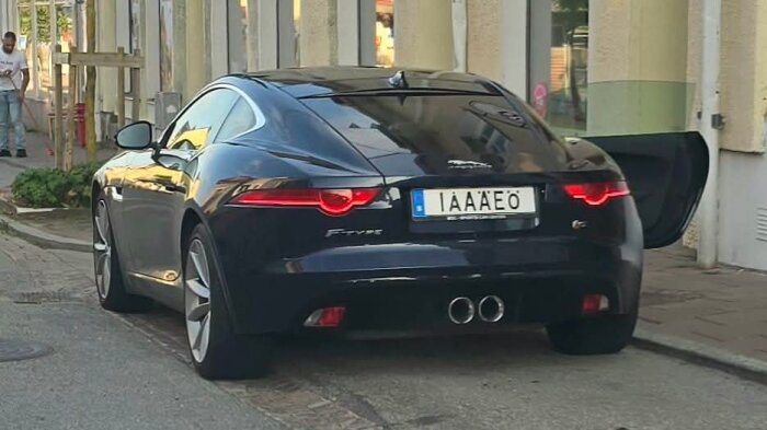 Svart sportbil parkerad vid trottoar med bakdörr öppen, registreringsskylt med texten "ÅIÅAEIÖ".