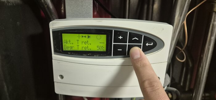 En person justerar inställningarna på en fjärrvärmecentral, modell ECL110, med en digital display som visar temperaturvärden.