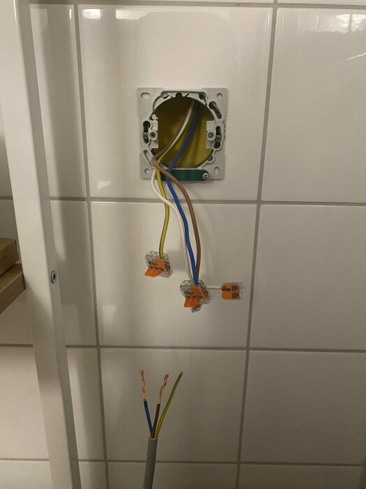 Elektriska kablar installerade i en väggdosa på en kaklad vägg, med lösa kabeländar och anslutningsklämmor.