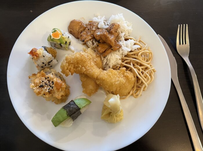 Tallrik med thaibuffé, sushi och dumpling, ovanpå ris och nudlar, serverad på ett mörkt bord bredvid gaffel och kniv.