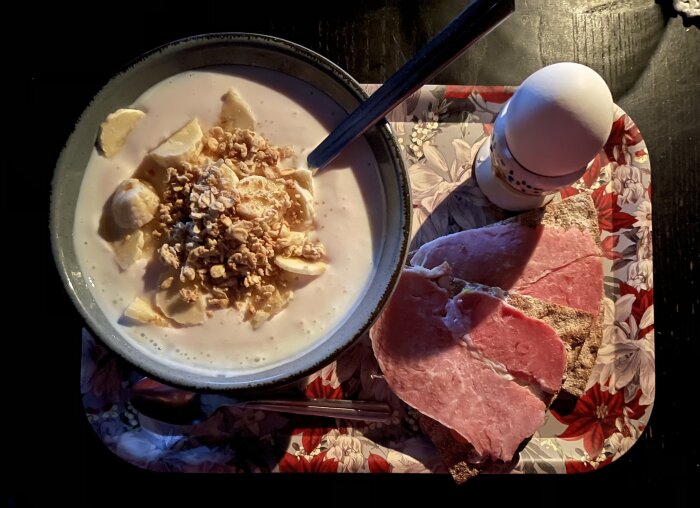 Frukost på en blommig bricka: skål med kvarg/fil, banan och granola, två fröknäcke med julskinka och ett kokt ägg i äggkopp.