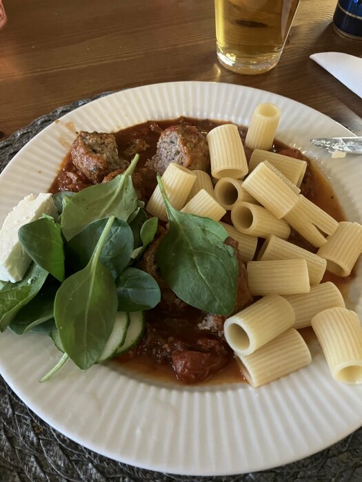 Tallrik med italienska köttbullar, rigatoni, grönt och fetaost, serverad med ett glas öl i bakgrunden.