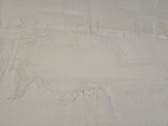 Ojämna ytmönster på en vägg täckt med microcement, synliga strukturer och färgskillnader efter bearbetning och slipning i ett badrumsprojekt.