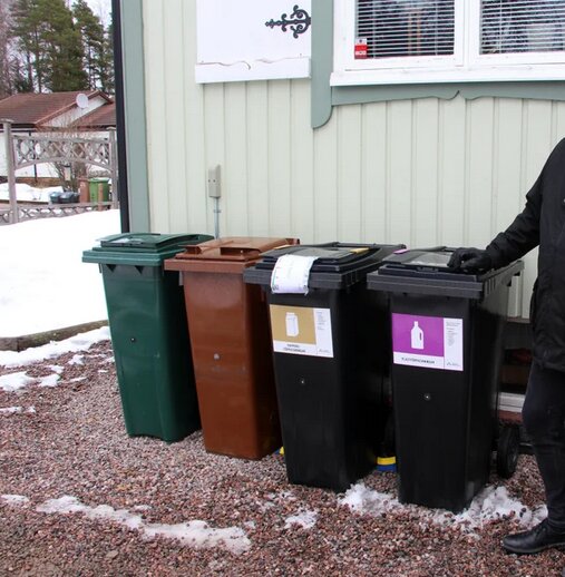 Fyra soptunnor i olika färger står på en grusgång vid ett hus, omgivna av snö.