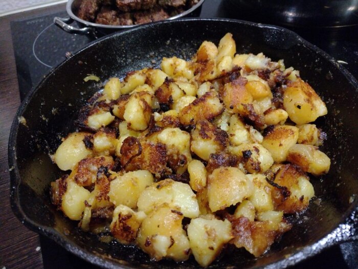 Stekt potatis i en svart stekpanna, serverad bredvid en skål med platta köttbullar på en spis.