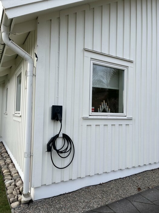 Vit yttervägg med elcentral och hängande kabel. Kabeln löper nedför väggen vid ett fönster, monterad längs en list. Provisoriska lösningar syns.