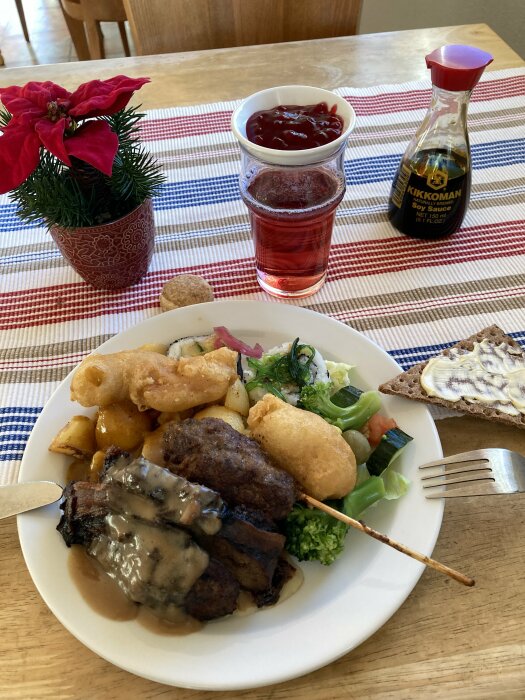 Tallrik med lunchbuffé med kött, potatis, sallad och bröd, tillsammans med ett glas röd dryck och en julblomma pÄ randig duk.