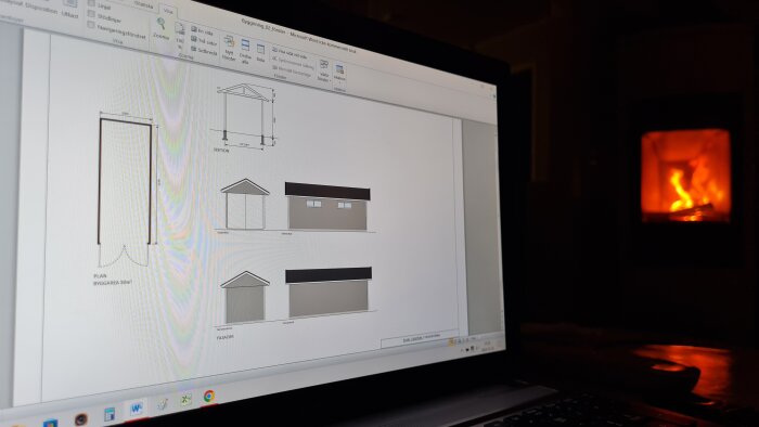 Dator med byggplaner på skärmen framför en tänd braskamin.