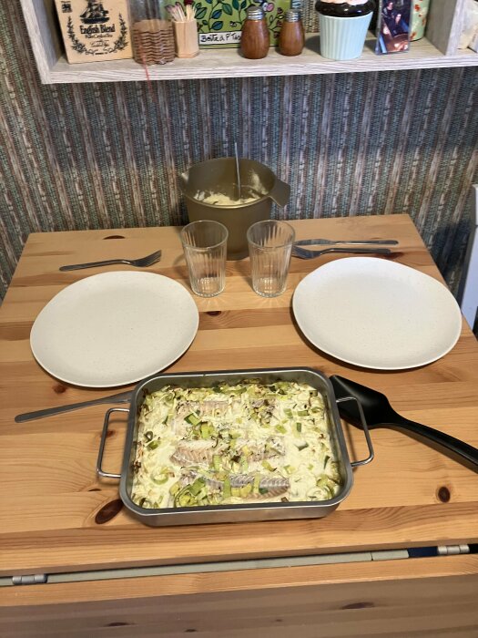 Ugnsform med tillagad torsk och spetskÄl pÄ trÀbord, omgiven av tvÄ tallrikar, bestick och glas.