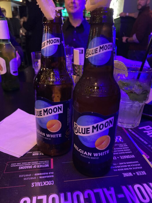 Två Blue Moon Belgian White-flaskor med citrusgarnering på en bardisk, omgivna av servetter och dryckesmeny i bakgrunden.