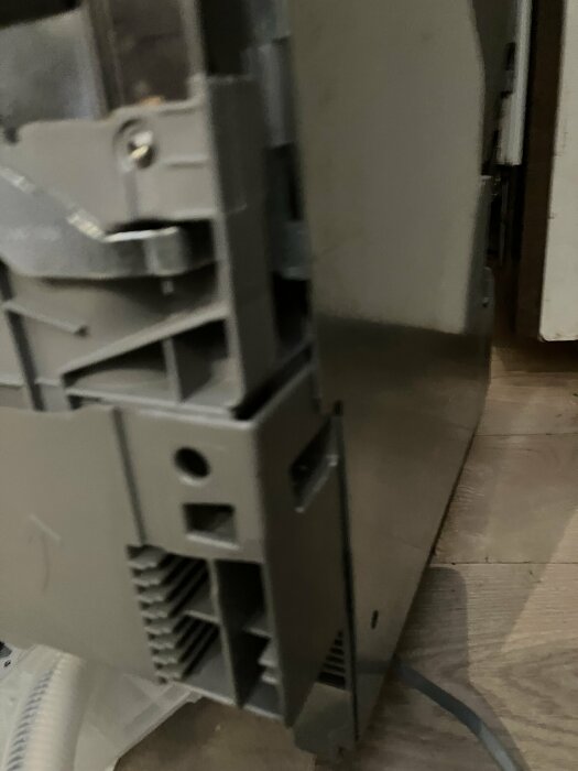 Grå hushållsmaskin med skruvar och plastdetaljer, avloppsslang skymtar i bakgrunden. Personen försöker få loss en lucka.