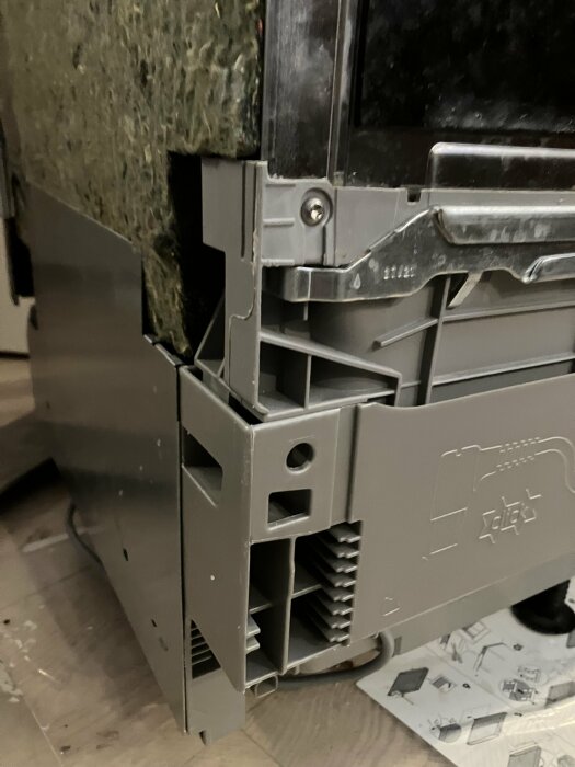 Närbild av en köksapparat med delvis öppet sidopanel som visar en skruv nära en mekanism. Apparaten står på ett trägolv.