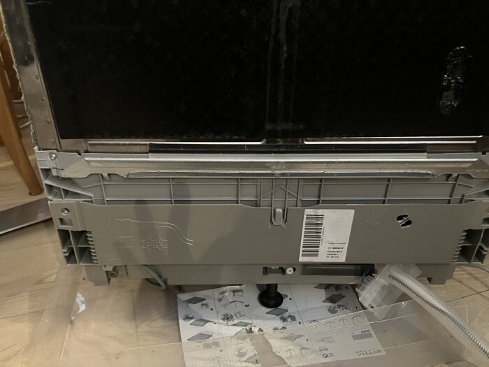 Baksida av diskmaskin där någon försöker nå avloppsslangen, instruktioner på golvet, svårt att avlägsna luckan.