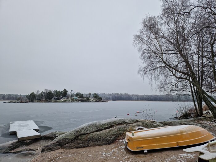 Isig sjö med en gul båt upp och ner, brygga och träd nära strandkanten, vinterlandskap med snötäckta klippor och skog i fjärran.