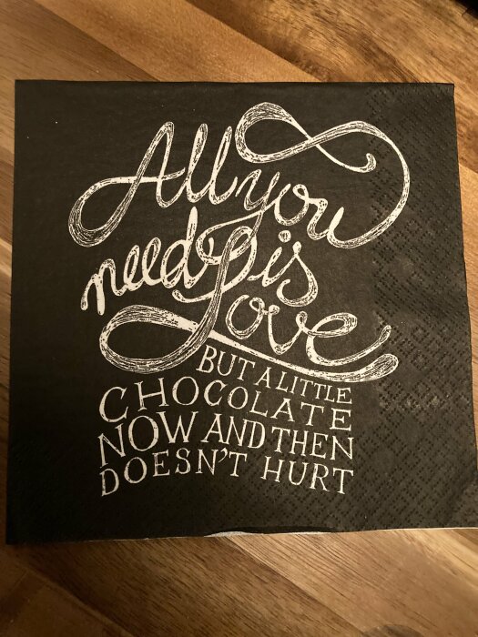 Svart servett med vit text: "All you need is love, but a little chocolate now and then doesn't hurt." Ligger på en träyta.