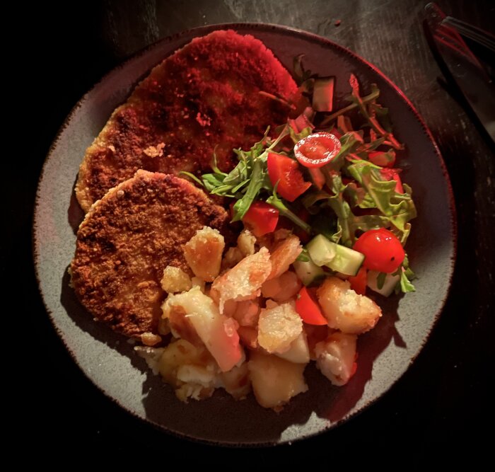 Tallrik med schnitzel, vitlöksstekt potatis och sallad med tomater och gurka under rött ljus.