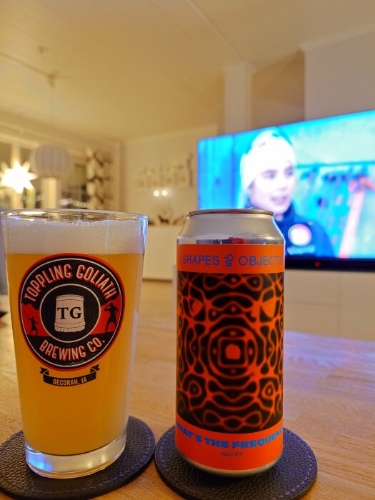 Ölglas med Toppling Goliath-logga bredvid en Shapes & Objects-burk i ett vardagsrum med TV i bakgrunden.