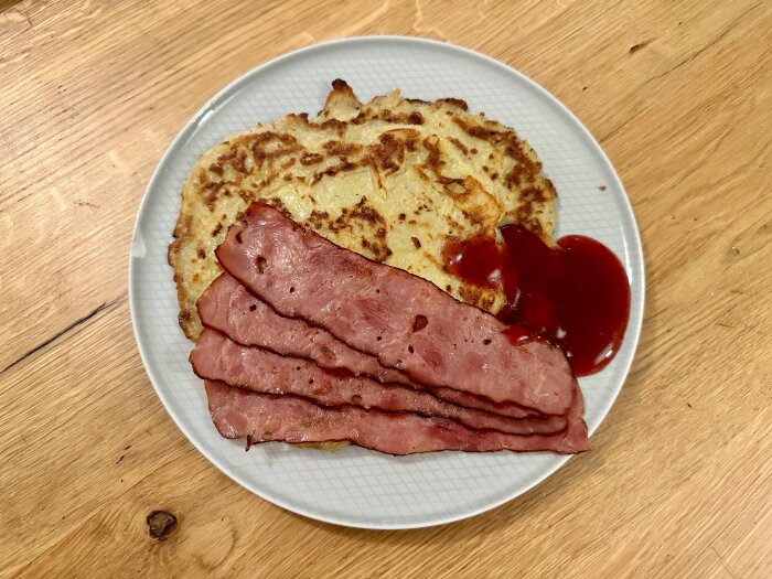 Raggmunk serverad med stekt bacon och lingonsylt på en vit tallrik, placerad på ett träbord.