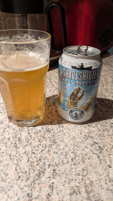 En ölburk med texten "Shapeshifter West Coast IPA" står bredvid ett glas halvfullt med öl på en köksbänk.