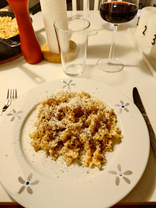 Pasta carbonara på tallrik med riven ost och peppar, glas vin och tom kryddkvarn i bakgrunden.