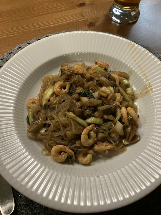 Wokade glasnudlar med räkor och grönsaker i en vit tallrik på ett bord.