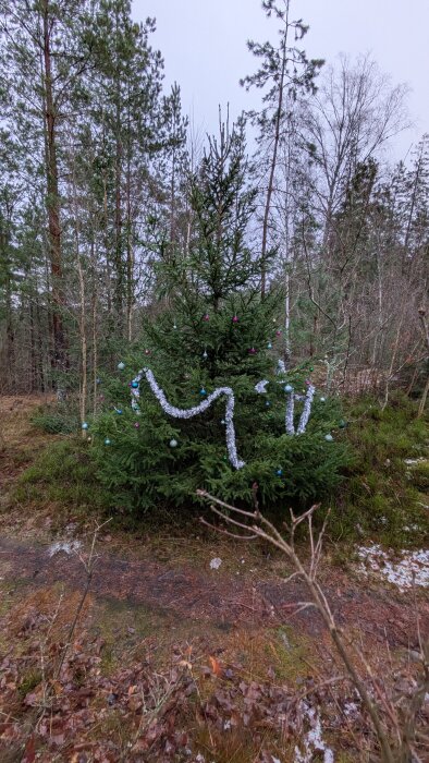 Grön gran dekorerad med blå och rosa kulor samt silverglitter i en skogsmiljö. Marken är täckt av löv och lite snö.