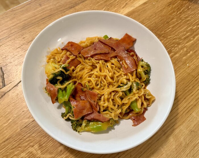 En tallrik med nudlar, skinka och broccoli, serverad på ett träbord.