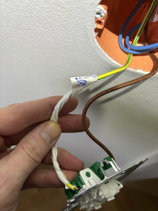 Gulgrön kabel märkt "lampa" syns kopplad till strömbrytare i en vit vägg; person håller brytaren, visar ovanlig användning av kabel.