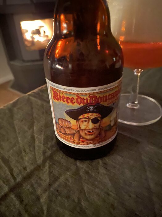 En flaska "Bière du Boucanier" med en etikett av en pirat, placerad nära ett vinglas och en brasa i bakgrunden.