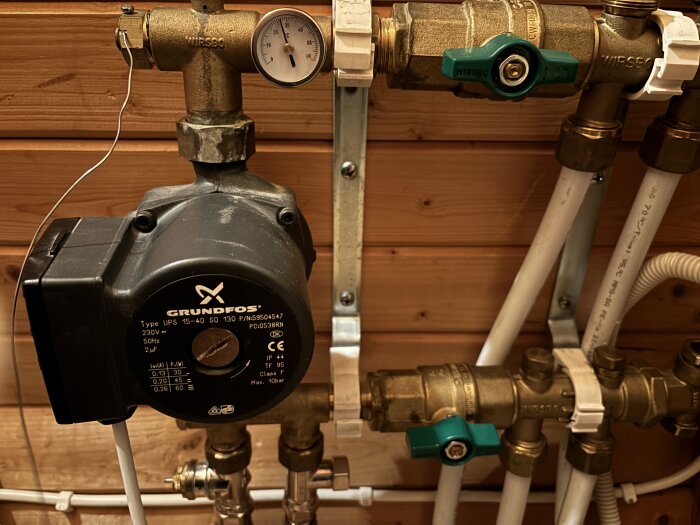 Pump och rörsystem för golvvärme, inklusive ventiler med gröna handtag och mätare, monterade på vägg. Grundfos-pumpen visas också.