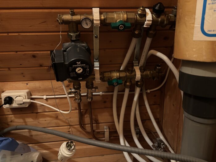 Golvvärmefördelare med ventiler och pump installerad på trävägg, synliga rör och elkontakt. Ventilerna är problematiska enligt inlägget.