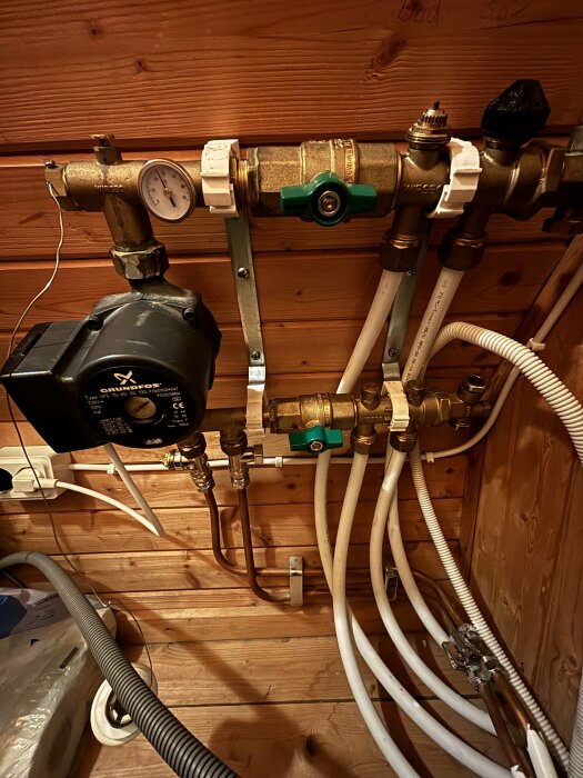 Golvvärmefördelare med rör och grön ventil i träbeklädd källare, plus tryckmätare och pump, relaterat till problem med golvvärmen.