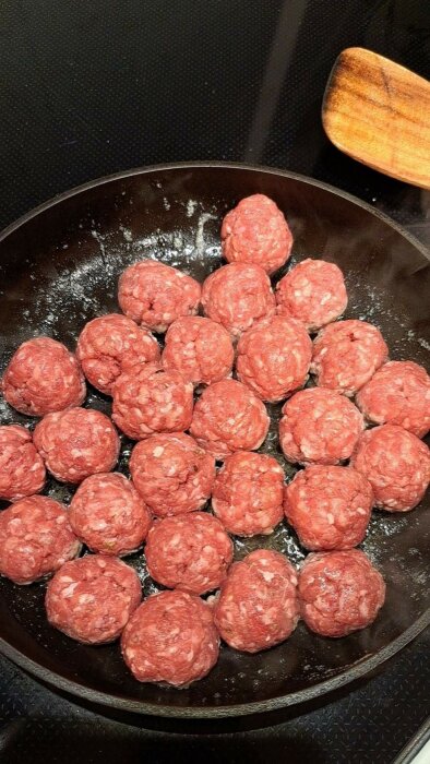 Råa köttbullar i en stekpanna med en träslev bredvid.