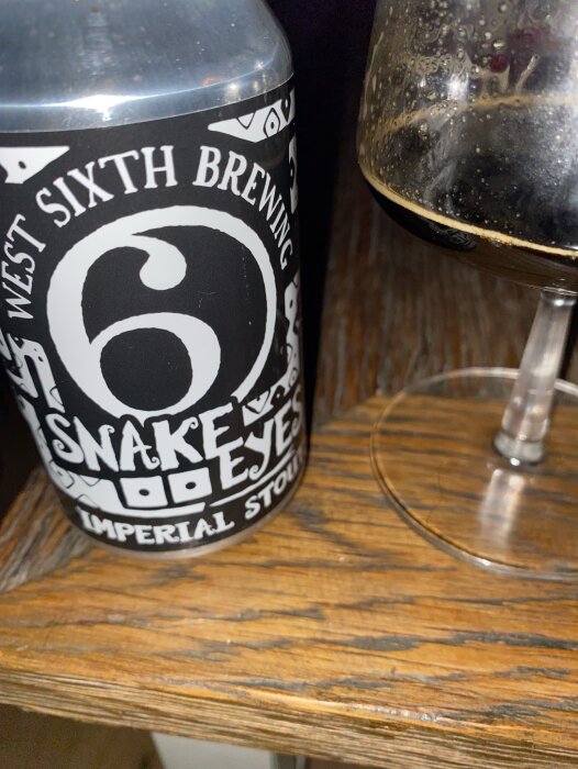 Burk med etikett "West Sixth Brewing Snake Eyes Imperial Stout" bredvid ett glas med mörkt öl på ett träbord.