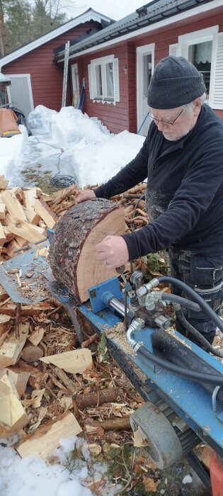 Man klyver ved med en vedklyv nära ett rött hus, omgiven av snötäckt mark och vedhögar.