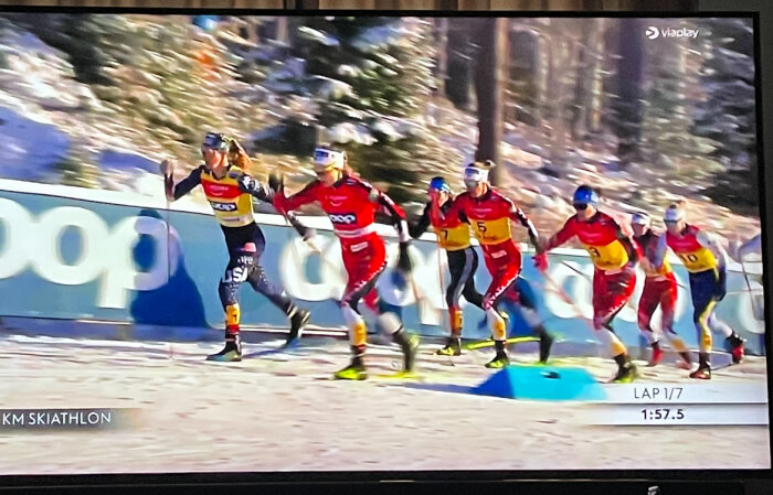 Damer tävlar i 20 km skiathlon, åker skidor snabbt i följd, omgivna av snötäckta träd och en reklamskylt i bakgrunden.