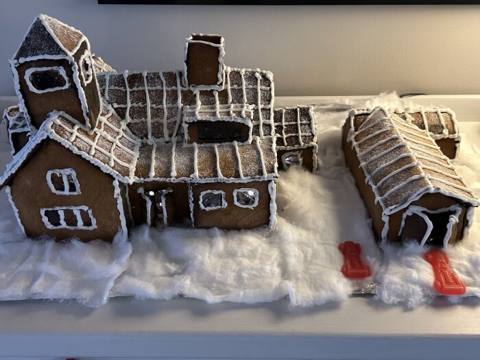 Pepparkakshus omgiven av bomullssnö, med detaljer av glasyr. Två röda godisbitar framför föreställer bilar.