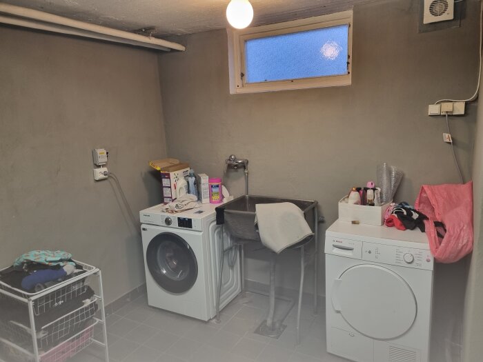 Tvättstuga med tvättmaskin, torktumlare och en vask. Flera eluttag syns på väggarna, samt förvaring av tvättmedel och en korg med kläder.